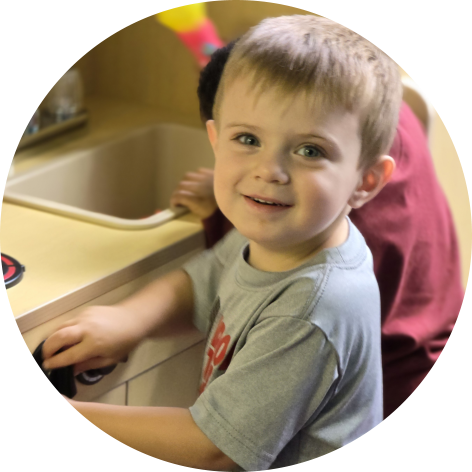 A young boy with blonde hair is standing at a play kitchen counter