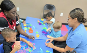 a group of four young children and two adults at a blue table, engaged in a play activity